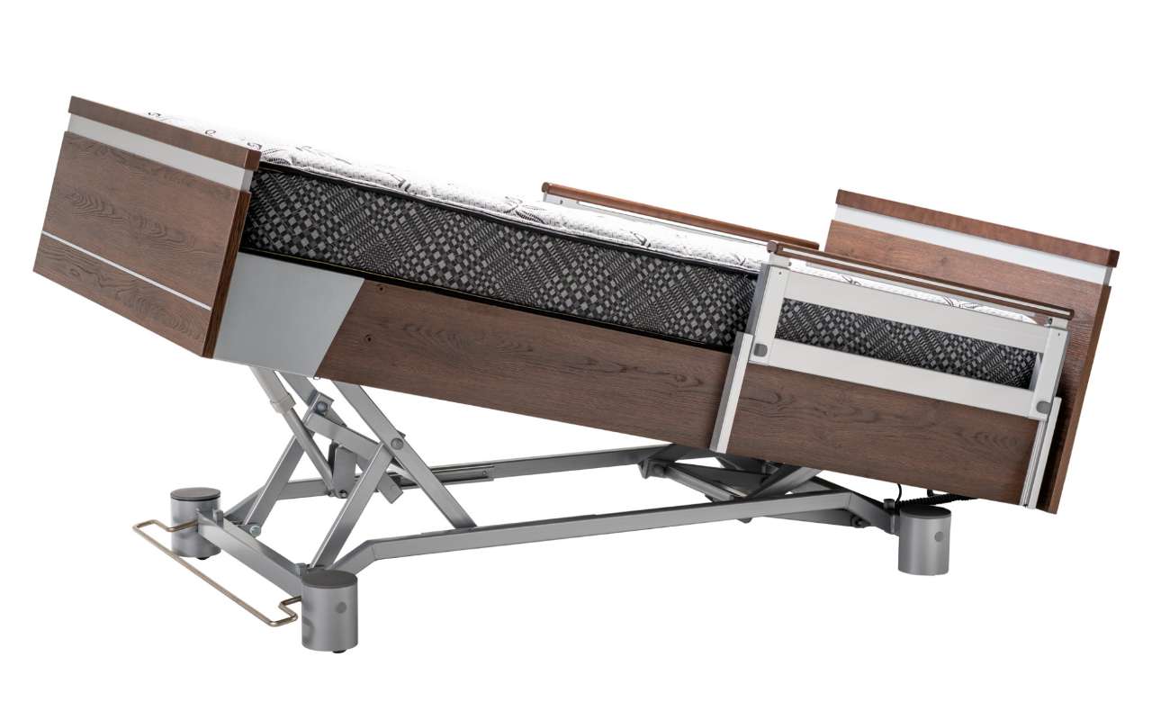 An adjustable hospital bed with wooden side rails reclines at an angle on a metal frame with a patterned mattress.