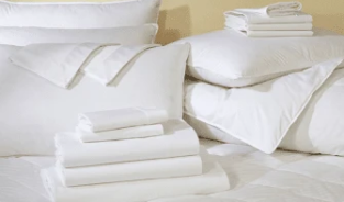 A neatly stacked set of white bed linens, including pillows, sheets, and folded blankets, on a bed.